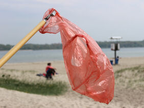 Eine weggeworfene Plastiktüte an einem Greifarm, im Hintergrund ein Gewässer