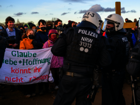 Zwei Polizisten unterhalten sich im Vordergrund während im Hintergrund demonstrierende ein Schild hochhalten, auf dem "Glaube Liebe + Hoffnung könnt ihr nicht räumen" steht. 