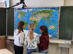Kinder schauen auf eine Weltkarte vor einer Tafel.