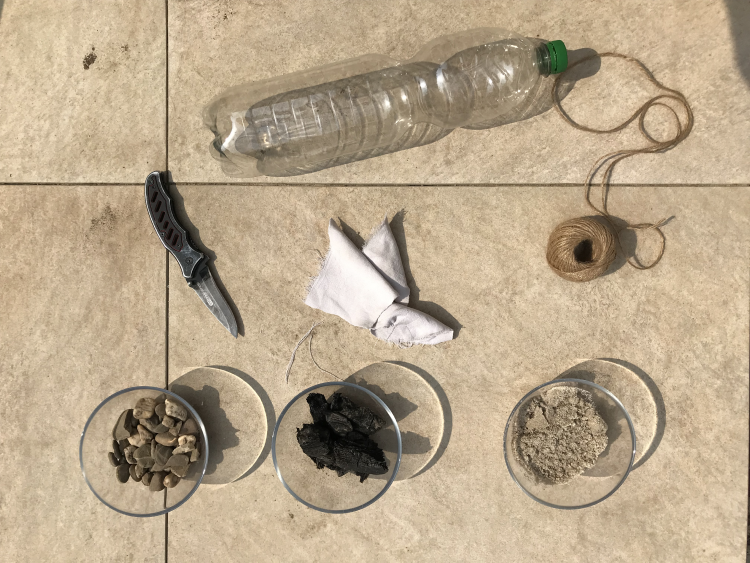 Das Bild ist eine Aufnahme von Materialien für den Wasserfilter: Messer, Plastikflasche, Kordel, Steine, Sand und ein Tuch.